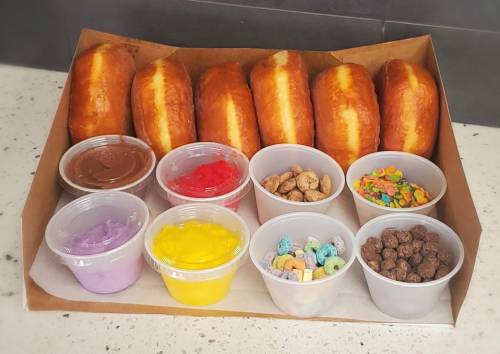 A tray of six donuts with colorful toppings and dips in cups, including chocolate, sprinkles, and frosting.