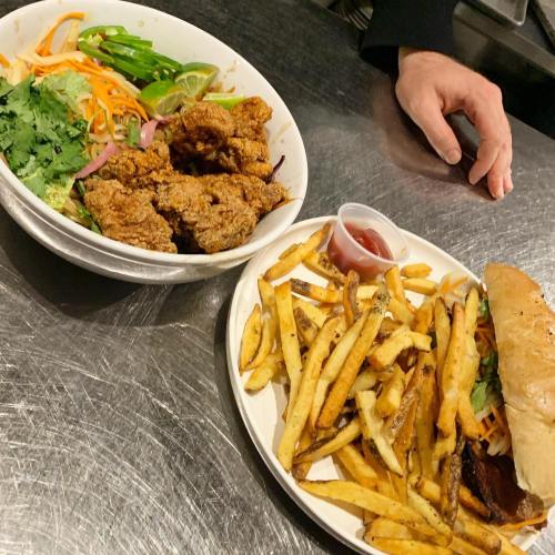 A bowl of fried chicken with salad and a plate of fries next to a sandwich on a metal surface.