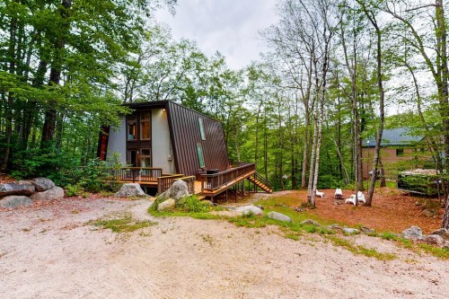 Modern cabin nestled in a wooded area, featuring a sloped roof and a wooden deck surrounded by trees and rocks.