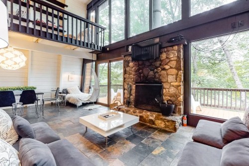 Cozy living room with stone fireplace, large windows, and modern furniture, surrounded by trees.