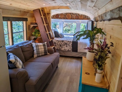 Cozy interior of a tiny home with a sofa, bed, and natural wood accents, surrounded by large windows and plants.