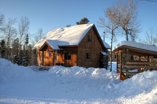 A wooden cabin with a snowy roof, surrounded by trees and a clear blue sky, in a winter landscape.