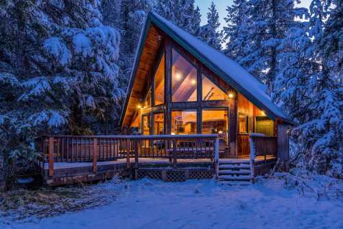 A cozy A-frame cabin surrounded by snow-covered trees, illuminated warmly at dusk.