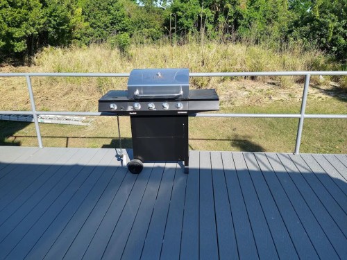 A black gas grill on a wooden deck, surrounded by greenery and open space in the background.