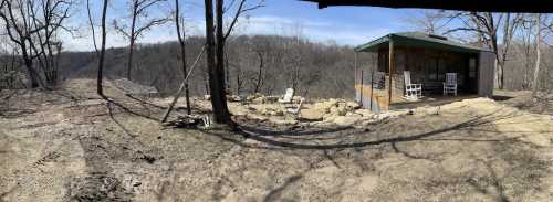 A rustic cabin surrounded by bare trees and a rocky landscape, with a view of rolling hills in the distance.