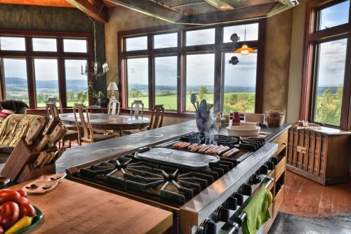 A spacious kitchen with a large stove, cooking sausages, and panoramic windows showcasing a scenic view.