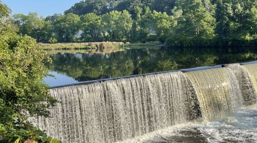 A serene view of a waterfall cascading over a dam, surrounded by lush greenery and a calm reflective lake.