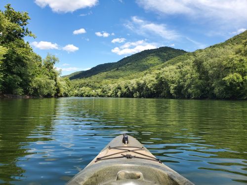 A kayak on calm water, surrounded by lush green trees and mountains under a blue sky with fluffy clouds.