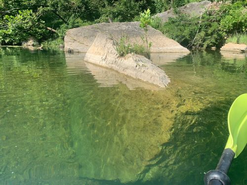 A clear, calm body of water with a large rock partially submerged and surrounded by greenery. A kayak paddle is visible.