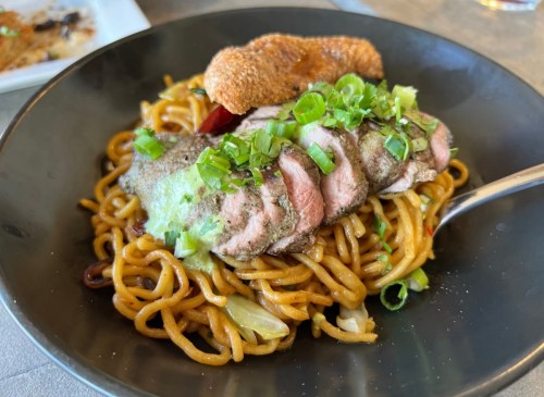 A bowl of stir-fried noodles topped with sliced beef, green onions, and a crispy garnish.