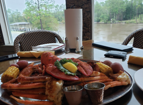 A seafood platter with crab, lobster, corn, and potatoes, set on a table with a view of a river and trees.