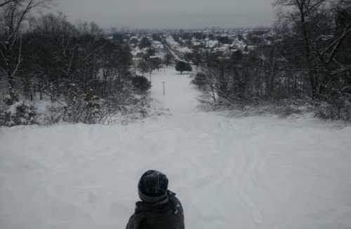 A child sits in the snow, looking down a snowy hill towards a quiet, snow-covered neighborhood.