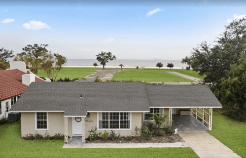 A single-story house with a gray roof, surrounded by green grass and trees, overlooking a body of water in the background.