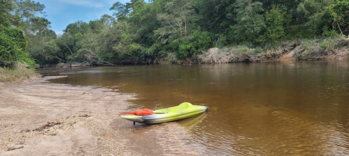 A calm river scene with a yellow kayak on the shore, surrounded by lush greenery and a clear blue sky.