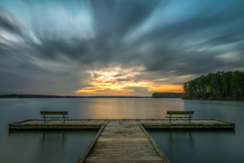 A serene lake view at sunset, featuring a wooden dock with two benches and dramatic clouds in the sky.