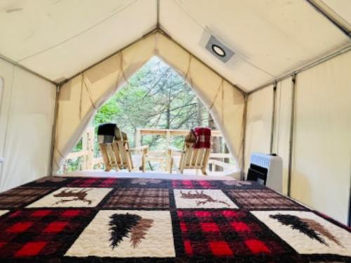 Interior of a tent with a quilted bedspread, looking out to a deck with two chairs and a forest view.