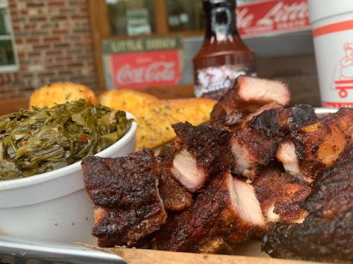 A plate of smoked barbecue meat, collard greens, and cornbread, with bottles of barbecue sauce in the background.