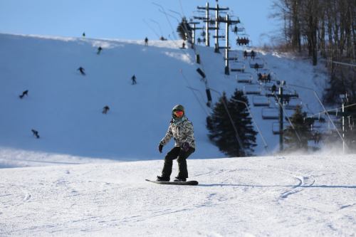 A child snowboards on a snowy slope with ski lifts and other skiers in the background under a clear blue sky.