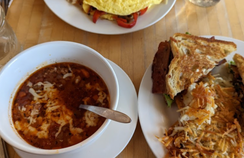 A bowl of chili topped with cheese, alongside a sandwich and a plate of hash browns with an omelet in the background.