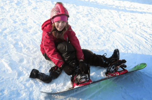 A smiling girl in a red jacket sits on snow, adjusting her snowboard while enjoying a winter day.