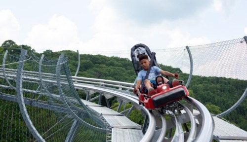 A child rides a red roller coaster on a winding track surrounded by green hills under a cloudy sky.