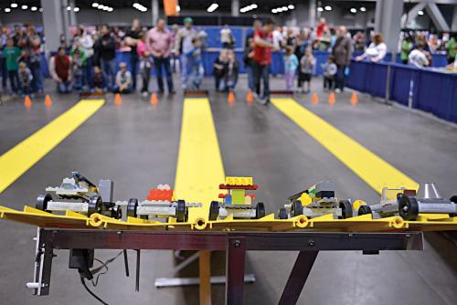 A view of a LEGO car racing track with colorful cars lined up, and a crowd watching in the background.