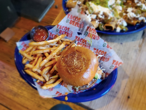 A plate with a burger on a bun, lettuce, and fries, served with a small cup of salsa. Tacos & Beers branding visible.