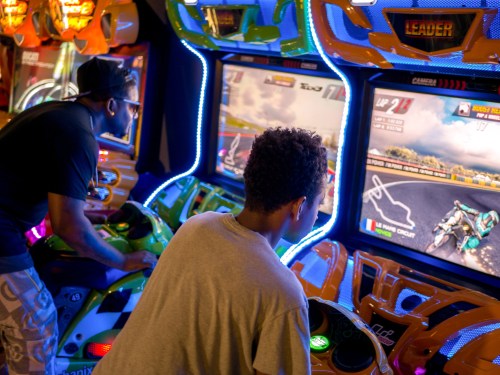 Two people playing motorcycle racing arcade games, focused on the screens with colorful lights around them.