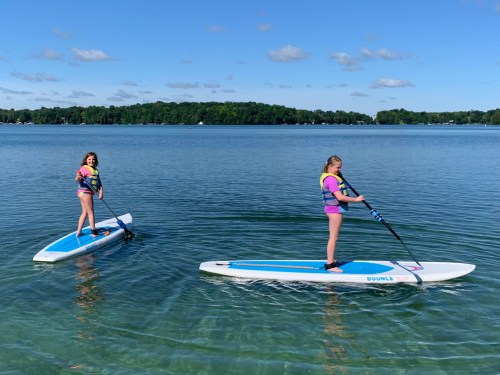 Two girls paddleboarding on a calm lake, surrounded by trees and a clear blue sky.