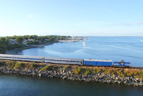 A train travels along a scenic waterfront, with boats docked and a clear blue sky in the background.