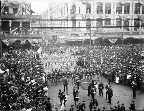 A historic parade scene with a large crowd, marching bands, and decorated buildings, capturing a festive atmosphere.