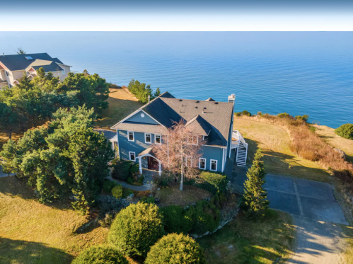 Aerial view of a blue house surrounded by greenery, overlooking a calm ocean under a clear sky.