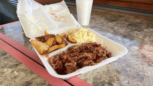 A takeout container with pulled pork, barbecue sauce, potato wedges, and creamy coleslaw, alongside a drink.