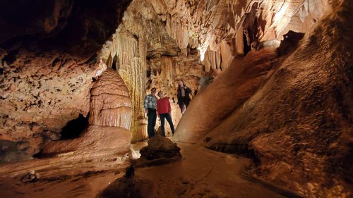 Two people stand in a cave surrounded by stalactites and stalagmites, with a stream flowing nearby.