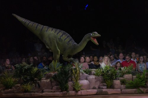 A dinosaur animatronic roars in front of an audience, surrounded by greenery in a dimly lit arena.