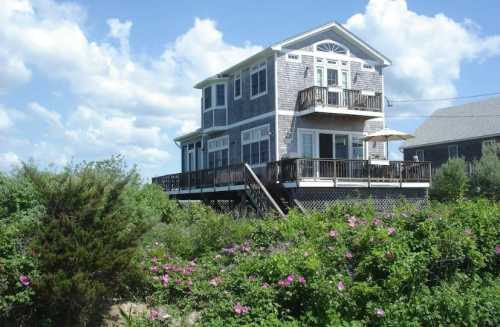 A two-story beach house surrounded by greenery and flowers, with balconies and a clear blue sky in the background.