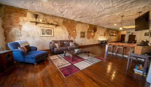 Spacious living room with exposed brick walls, wooden floors, a blue chair, and a coffee table on a patterned rug.