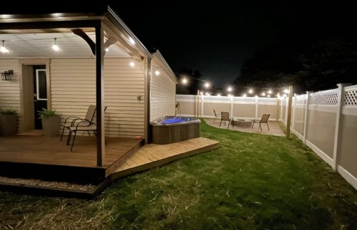 A backyard at night featuring a hot tub, patio chairs, and string lights illuminating the space.