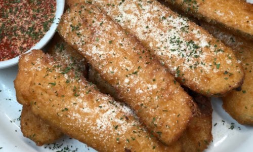 A plate of golden-brown fried zucchini sticks, sprinkled with parmesan and served with marinara sauce on the side.