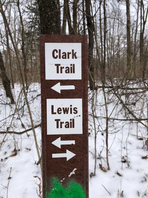 Trail sign indicating directions for Clark Trail and Lewis Trail, surrounded by bare trees and snow.