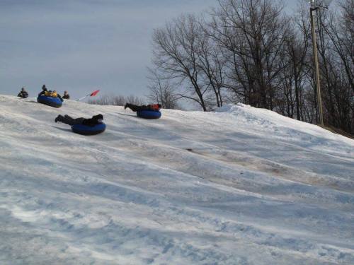 People sledding down a snowy hill on inflatable tubes, surrounded by trees and a cloudy sky.