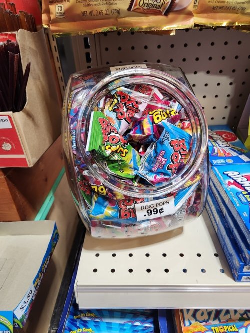A clear jar filled with colorful candy, including Ring Pops, displayed on a store shelf. Price tag shows 99 cents.