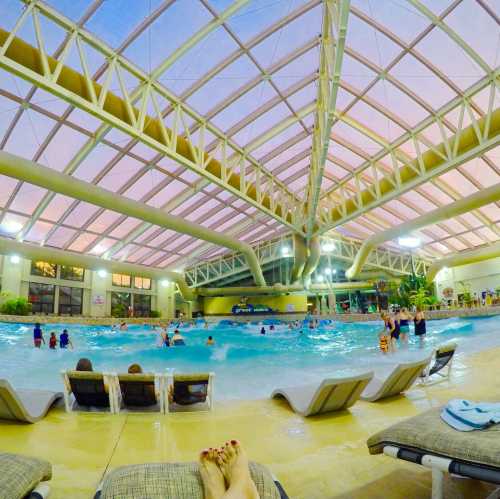 Indoor water park scene with a wave pool, people swimming, and lounge chairs under a large glass roof.