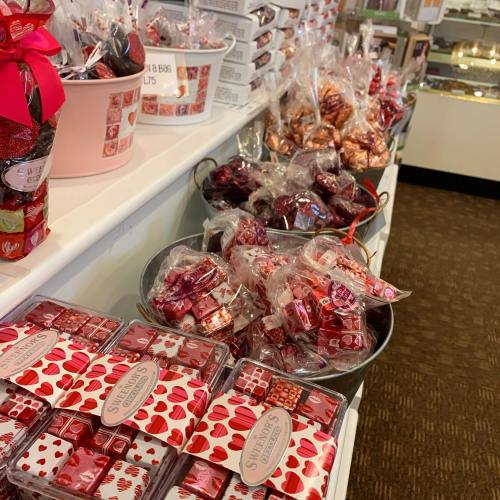 A display of assorted candies in bags and boxes, featuring red and pink packaging, arranged on a store shelf.