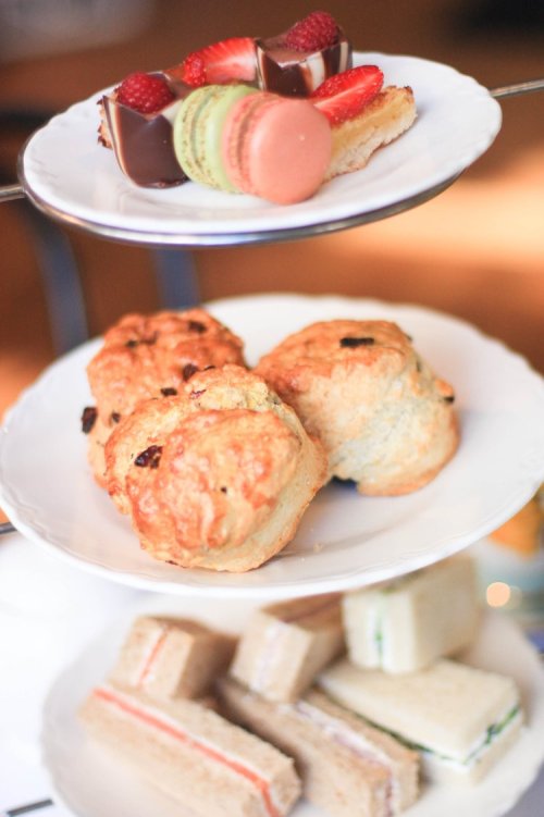 A tiered stand with assorted pastries: macarons, chocolate-covered treats, scones, and finger sandwiches.
