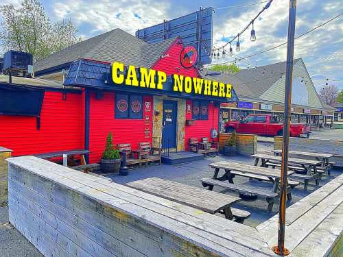 Bright red building with "CAMP NOWHERE" sign, outdoor seating area, and a cloudy sky in the background.