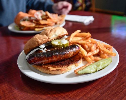 A close-up of a sausage sandwich with pickles and onions, served with curly fries on a plate.