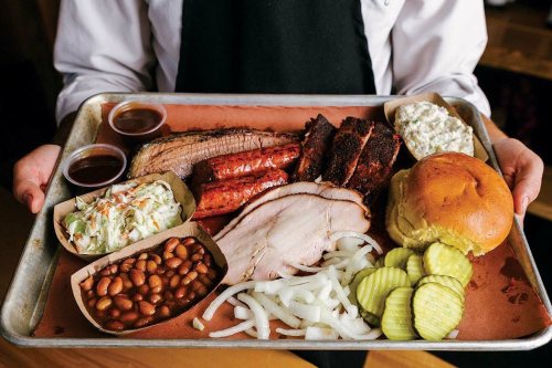 A metal tray filled with barbecue meats, sides of coleslaw, baked beans, pickles, onions, and a bun.