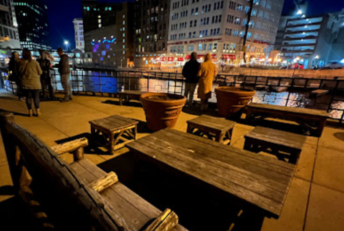 Nighttime view of a riverside area with wooden benches, large pots, and people socializing in the background.