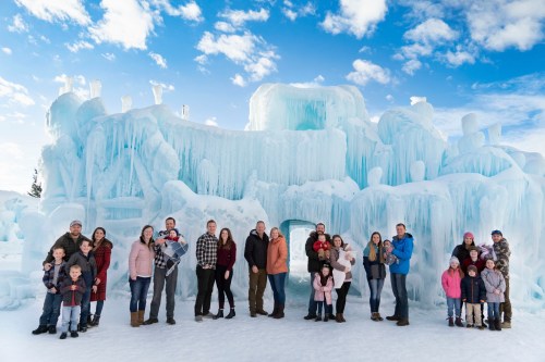 A large group of people poses in front of an ice sculpture under a blue sky with fluffy clouds.